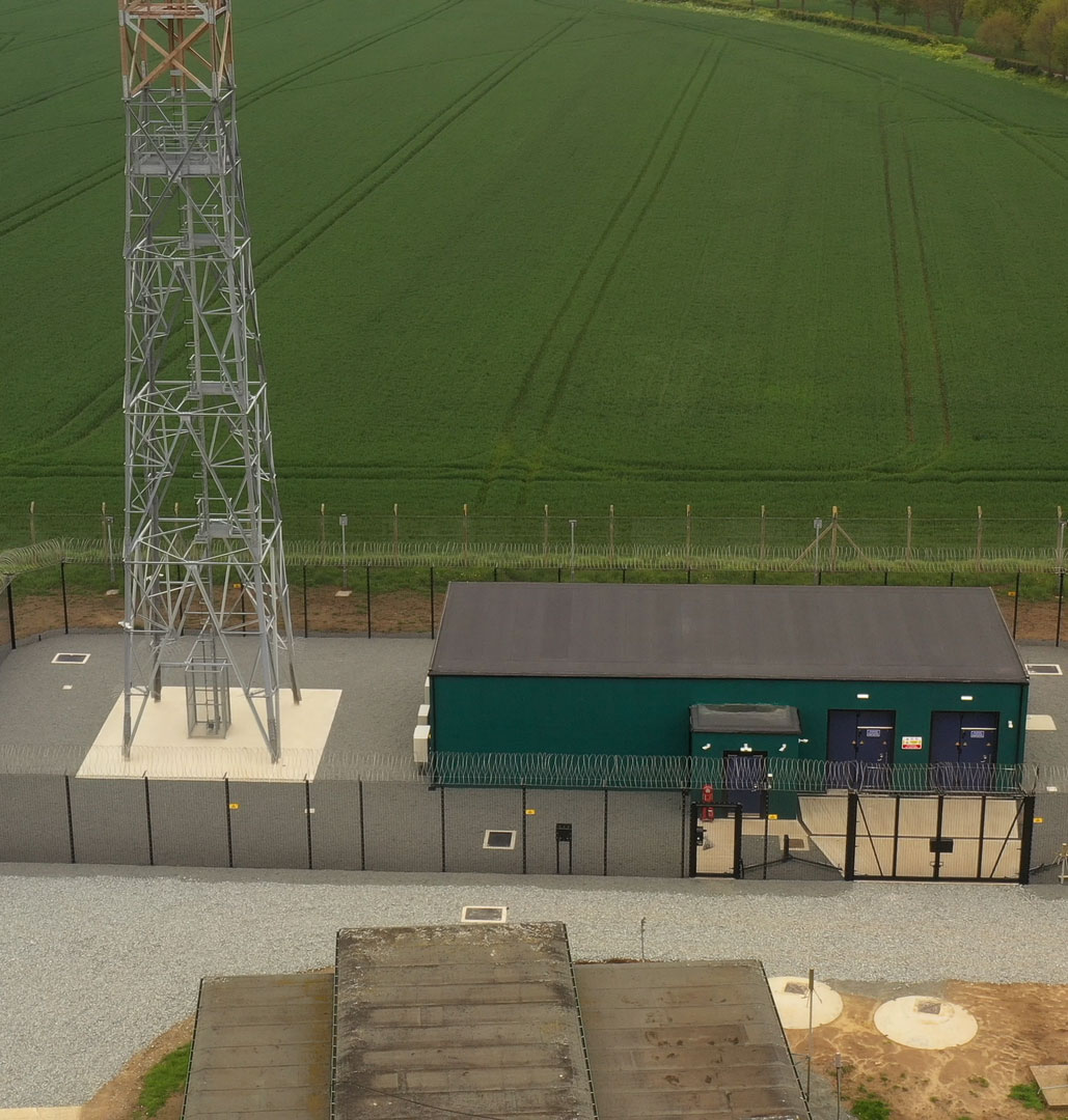 Aerial view of a perimeter security fence enclosing a remote FCDO communications mast and equipment building in a rural field