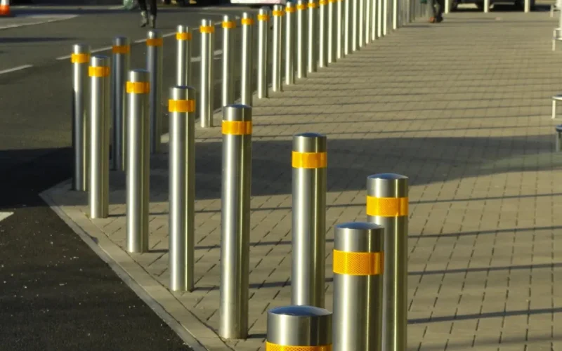 Row of shallow-mount stainless steel security bollards with yellow reflective bands along a pedestrian pavement